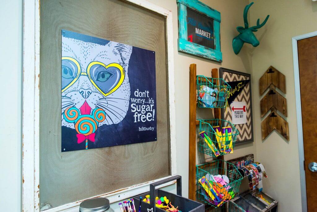 activities center rack with teal colored hanging wire baskets, animated character art of a cat with heart shaped lenses yellow glasses frame licking a teal and orange swirled design large flat lollipop saying: don't worry it's sugar free, a deer head in teal color, and a few other original art designs wall hanging
