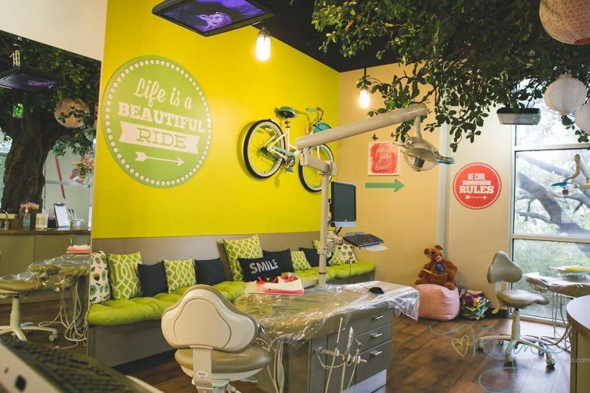 Hill Country Pediatric Dentistry and orthodontics office treatment area under a big tree with white and beige Chinese lamps balls view of the big real pair of bicycle hanging on the bright yellow wall along with a round retro style lime green colored sign with dotted frame in white and an arrow pointing right, saying: Life is a Beautiful Ride written in white positioned above a long seating bench covered with lime green base cushion and back cushions in lime green, black and white variation, a few other office signs in red and white visible on adjacent wall