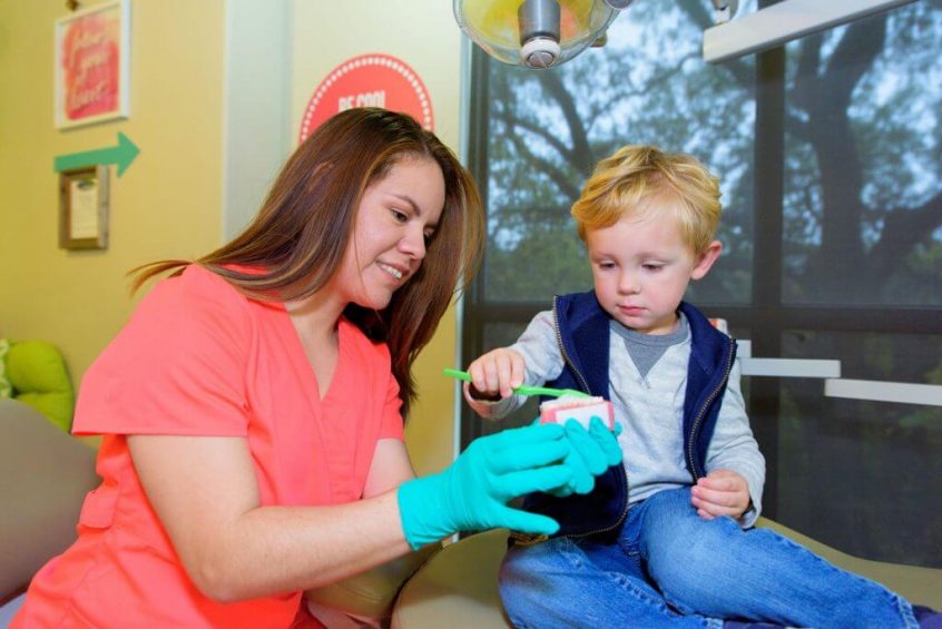 A young patient wearing blue jeans a gray sweatshirt and a blue open zippered vest holding a large sized green tooth brush is brushing the teeth of a gum and teeth model held by a team members of Hill Country Pediatric Dentistry and orthodontics office wearing orange office uniform and green exam gloves sitting next to the treatment bed on which the blond hair boy is sitting, the view of a few retro style red background with white writing office decor signs are visible on the wall behind them
