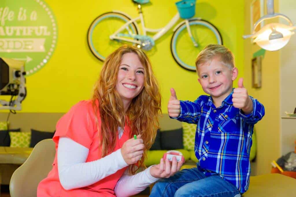 A young patient wearing blue jeans and a blue plaid flannel shirt signaling holding both his thumbs up sitting on a treatment bed and a smiling team members of Hill Country Pediatric Dentistry and orthodontics office wearing orange office uniform holding a large sized green tooth brush and a gum and teeth model sitting next to the treatment bed on which the boy is, view of the big real pair of bicycle hanging on the bright yellow wall along with a round retro style lime green colored sign with dotted frame in white and an arrow pointing right, saying: Life is a Beautiful Ride written in white positioned above a long seating bench covered with lime green base cushion and back cushions in lime green, black and white varied patterns on the wall behind them