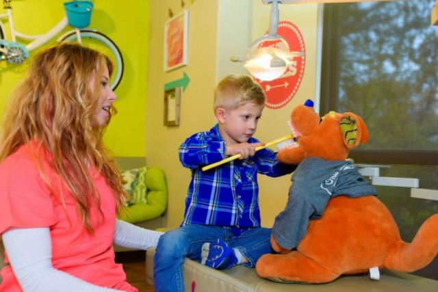 A young patient wearing blue jeans and a blue plaid flannel shirt holding a huge sized yellow tooth brush to brush the teeth of a child size office orange kangaroo stuffed figure who is wearing a gray tee shirt while both are sitting on the treatment bed with one of the team members of Hill Country Pediatric Dentistry and orthodontics office is supervising the boy, the view of the yellow bright wall behind her with part of the bicycle hanging from the wall is visible and a few retro style red background with white writing office decor signs are visible on the other wall