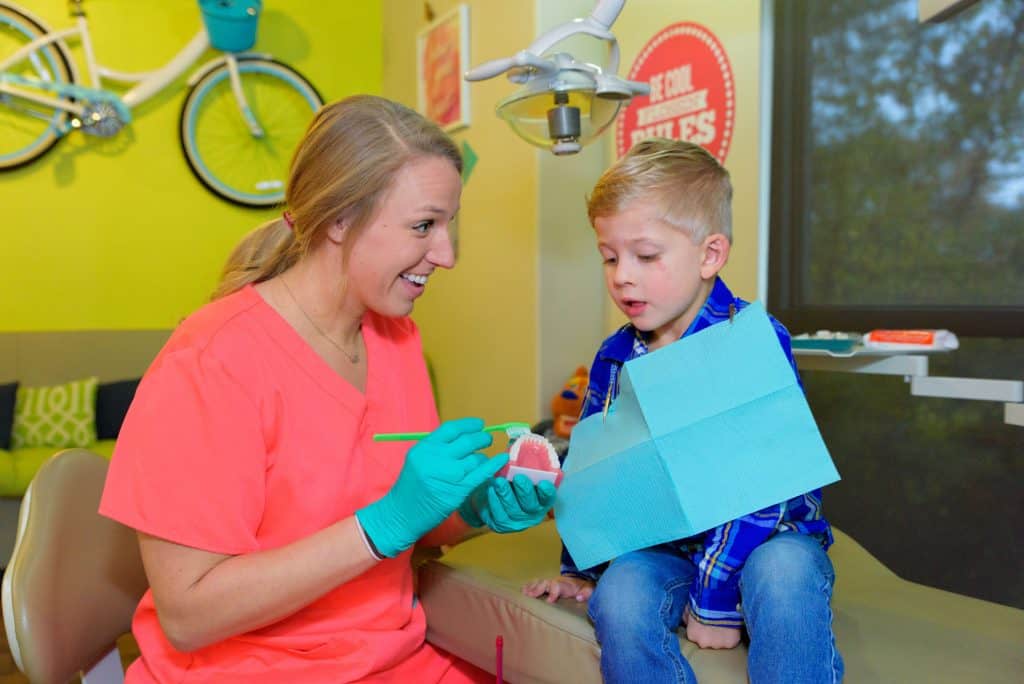 A young patient wearing blue jeans and a blue plaid flannel shirt and having a disposable napkin protection covering his chest sitting on the treatment bed with one of the team members of Hill Country Pediatric Dentistry and orthodontics office is smiling while sitting next to the treatment bed in the doctor chair wearing orange uniforms and green exam gloves on her hands holding a model of gum and teeth in one hand and a large green toothbrush in the other hand demonstrating to the young boy the proper procedure for effective teeth brushing, the boy is attentively looking at the model and toothbrush and interested in the educating demonstration, the view of the yellow bright wall behind her with part of the bicycle hanging from the wall is visible and a few retro style red background with white writing office decor signs are visible on the other wall 